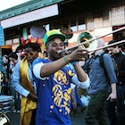 A young man with a bright green baseball hat and blue t-shirt plays a trombone in the street with other revellers, some donning Venetian masks, others feathers in their hair