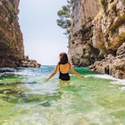 Young adult woman in black swimsuit walking into sea between rocks cliff in Croatia