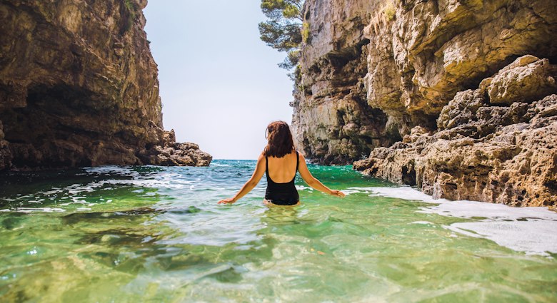 Young adult woman in black swimsuit walking into sea between rocks cliff in Croatia