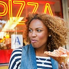 A close up of a black woman eating a slice of pizza outside a pizza restaurant in NYC