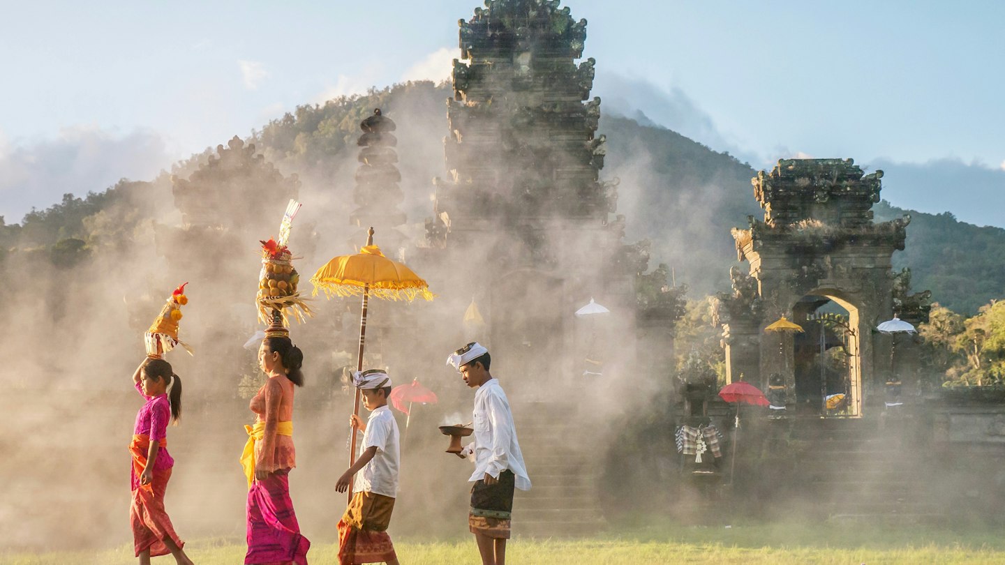 A mother and a teenaged girl are dressed in brightly colored sarongs, blouses, and sashes and are balancing tall fruit baskets on their heads. Two sons are dressed in sarongs and white shirts. The family is walking in front of an old stone temple building which has a smoky atmosphere.
938016384
religious