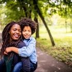 A mother is carrying her child on her back in the park as they both smile at something past the camera