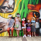 Two adults and three children stand in front of a wall covered in street art. The man holds a small cloud-shaped sign that covers his face and reads 'I heart graffiti'. The woman holds a similar sign that reads 'Art is not a crime'. The children hold similar sings, including ones that say 'Wow' and 'Cool'.© Heather Mason / Lonely Planet