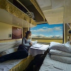 Young woman lies on the bed and looks through the window of the sleeper train crossing picturesque Tibet. Female tourist observing the landscape while traveling along the Trans-Himalayan railway.